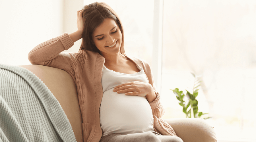 A lady on couch smiling holding her hand on her tummy where she is pregnant with a baby bub to be