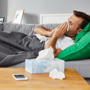 A man on couch laying down with his hands over his nose and a tissue to blow over his nostrils surrounded by other tissues used with his unwell cold
