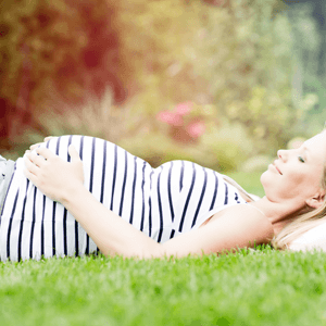 A woman laying on grass with a pillow and holding her arm at her stomach whre she is pregnant with a baby